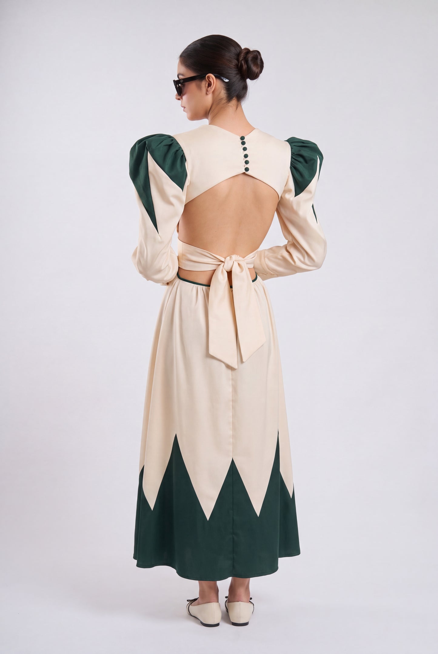 A woman shown from the back wearing a long cream dress with dark green contrast panels, puffed long sleeves, and a tied open back, photographed against a light gray studio background.