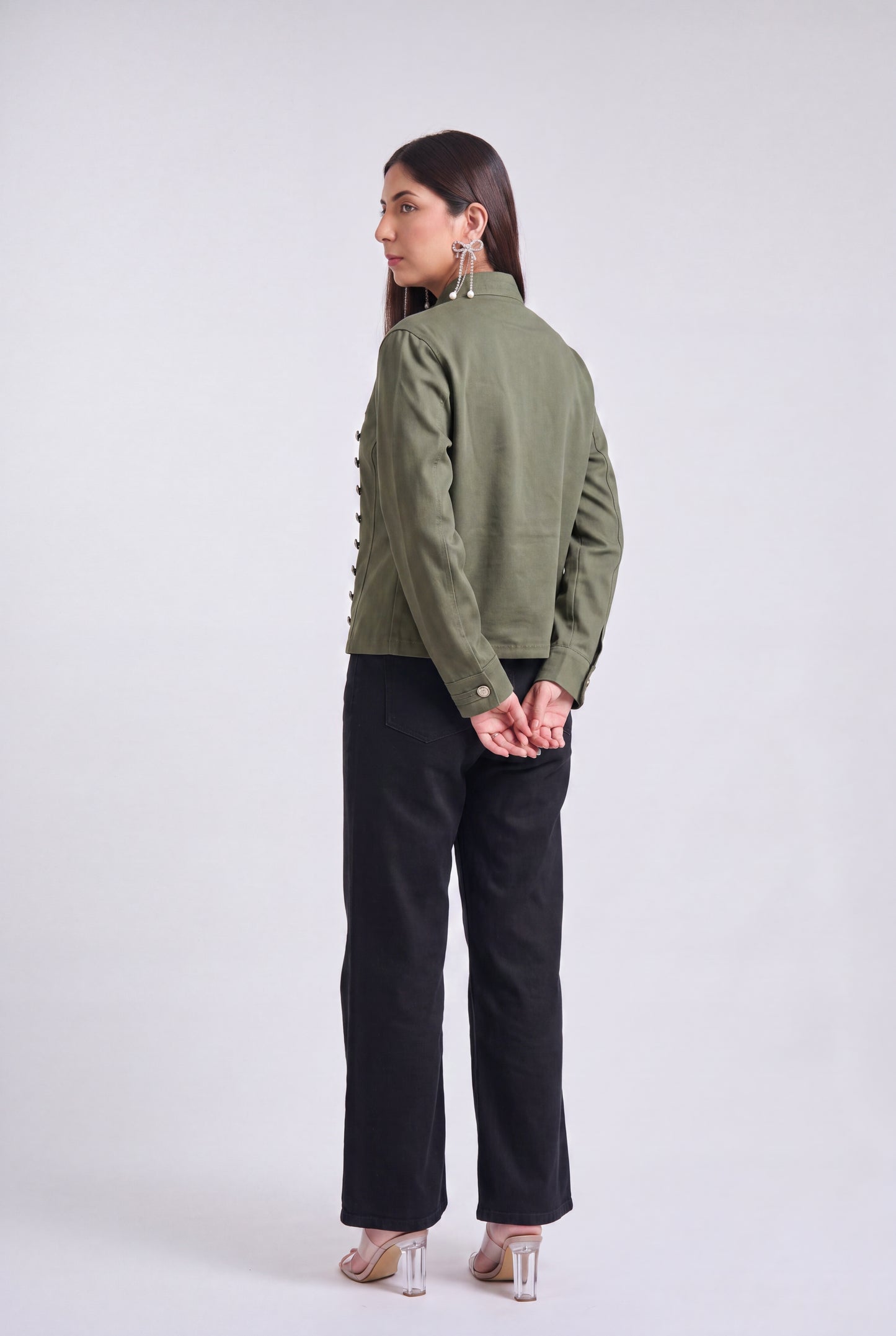 A woman modeling an olive green military-style jacket with silver buttons, paired with black wide-leg pants and clear high-heeled sandals, against a light gray studio background.