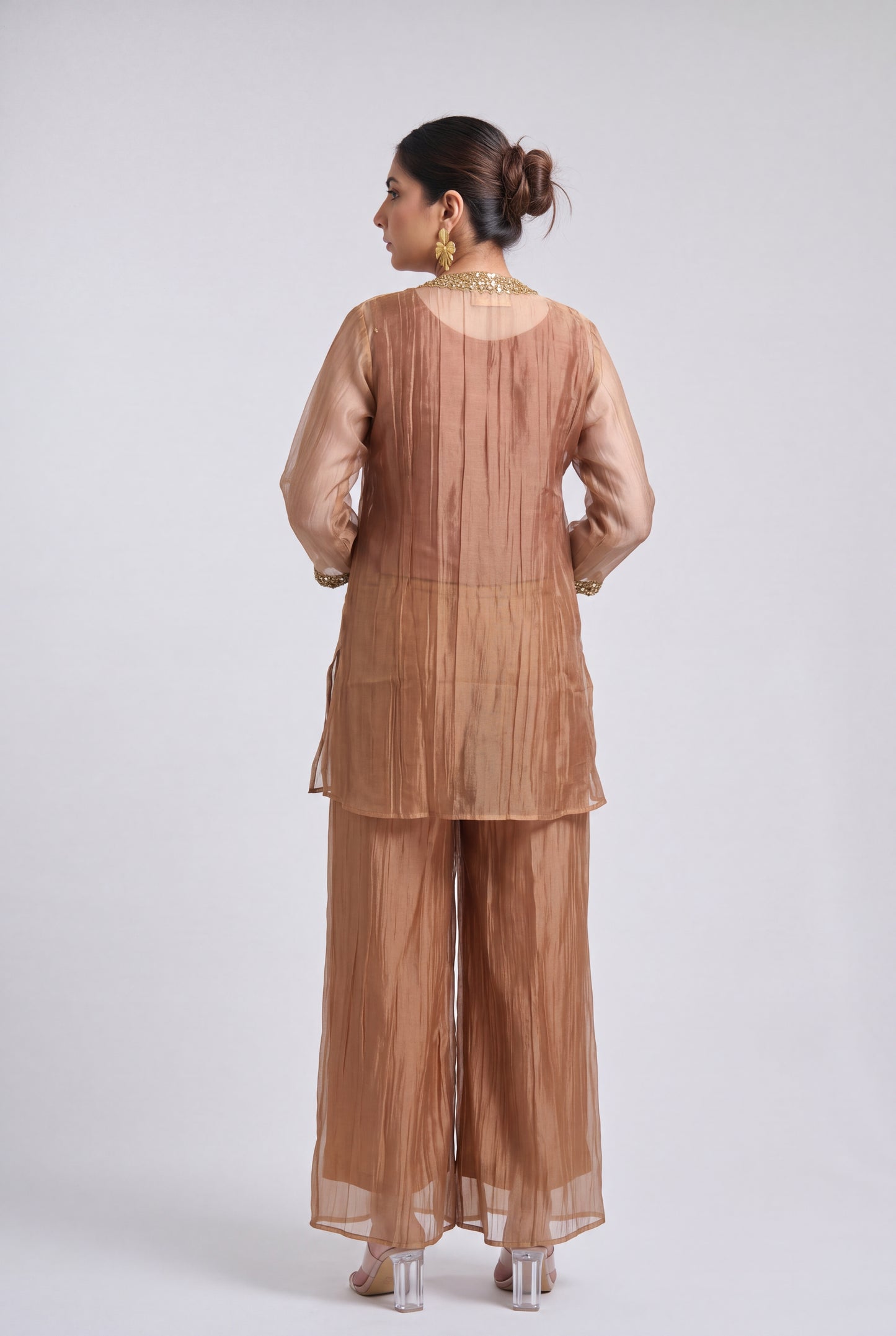 A woman is shown from the back wearing a matching two-piece traditional outfit in a warm taupe-brown color. The set features a long-sleeved tunic with a pleated back and wide-leg trousers, both made of a sheer, lightweight fabric.