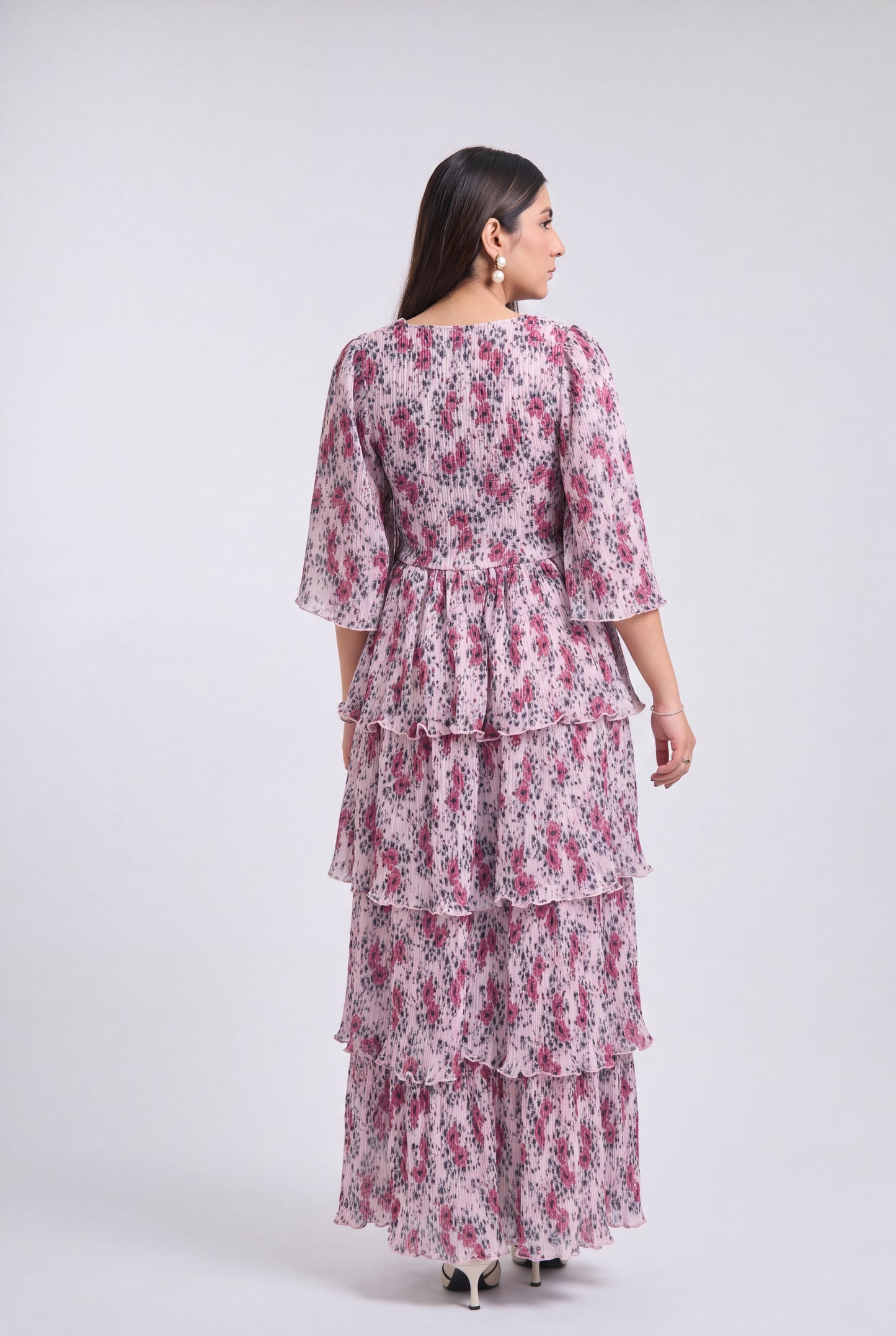 A woman shown from the back wearing a tiered maxi dress with a pink and purple floral print on a white base, photographed against a light gray studio background.