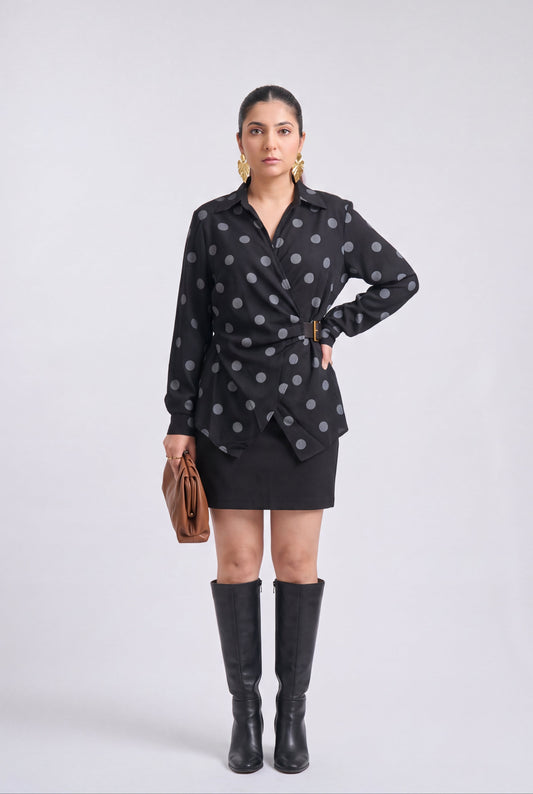 White and black polka dot wrap shirt with collared neckline, full sleeves, waist belt and asymmetrical hem, styled with a black mini skirt and knee-high boots.