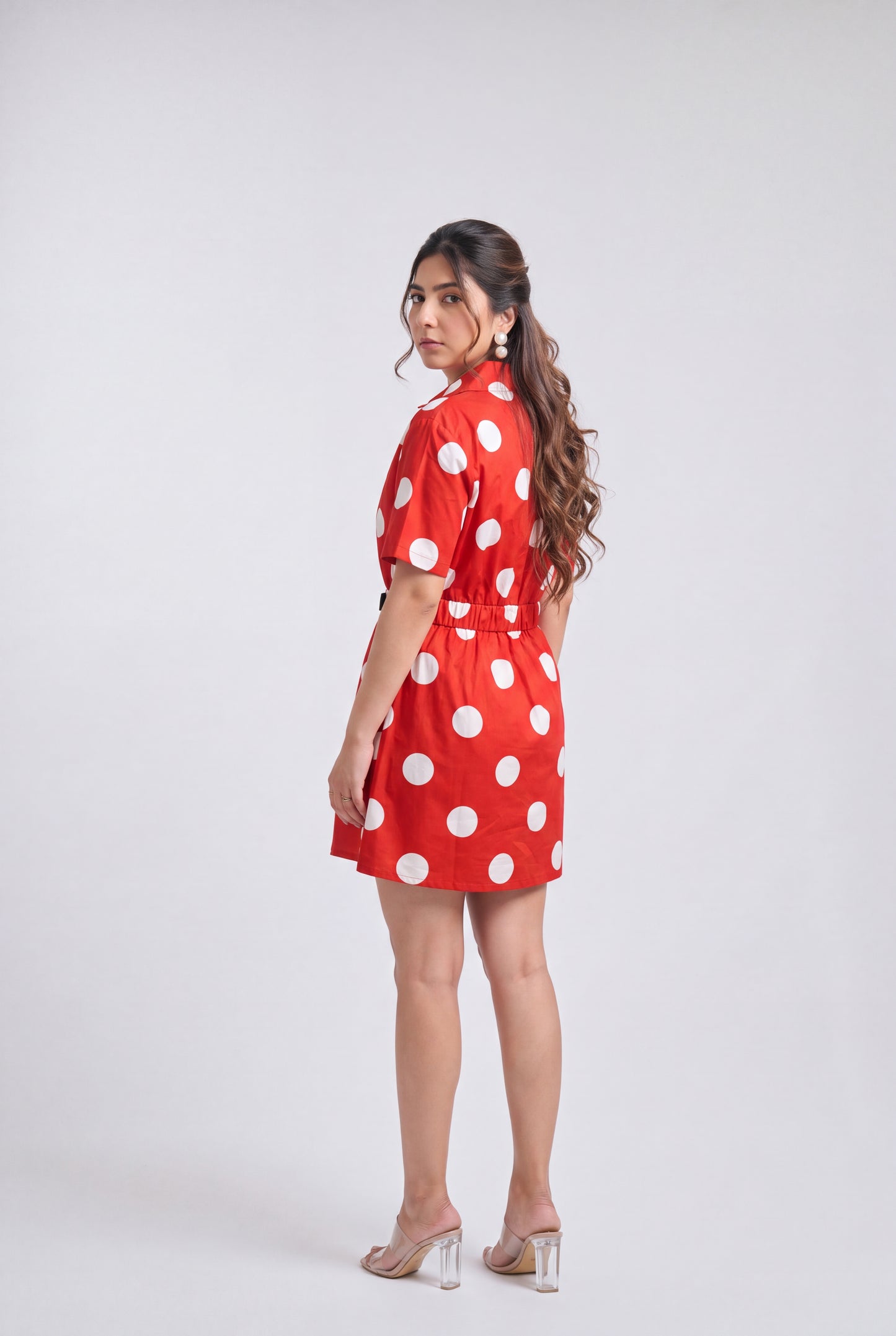 A woman modeling a red short-sleeve mini dress with large white polka dots, shown from a side-back angle against a light gray studio background.