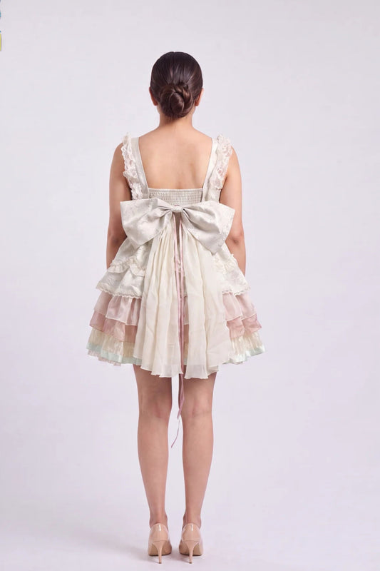 A model shown from the back wearing a short cream-colored dress with a large bow at the back, lace straps, and a tiered ruffled skirt, paired with beige heels against a light gray studio background.