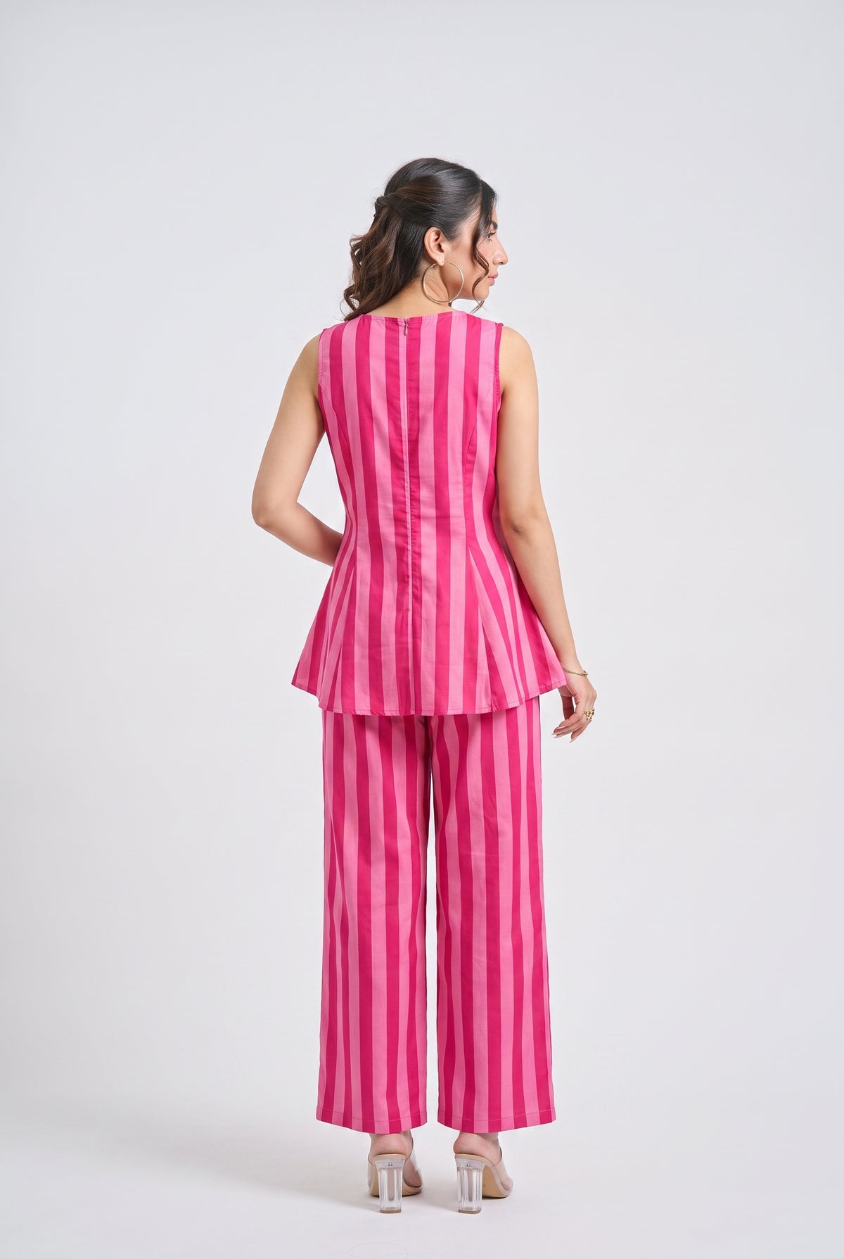 A woman is shown from the back wearing a bright pink and white vertically striped sleeveless peplum top and matching wide-leg trousers, paired with clear high-heeled sandals, against a light gray studio background.