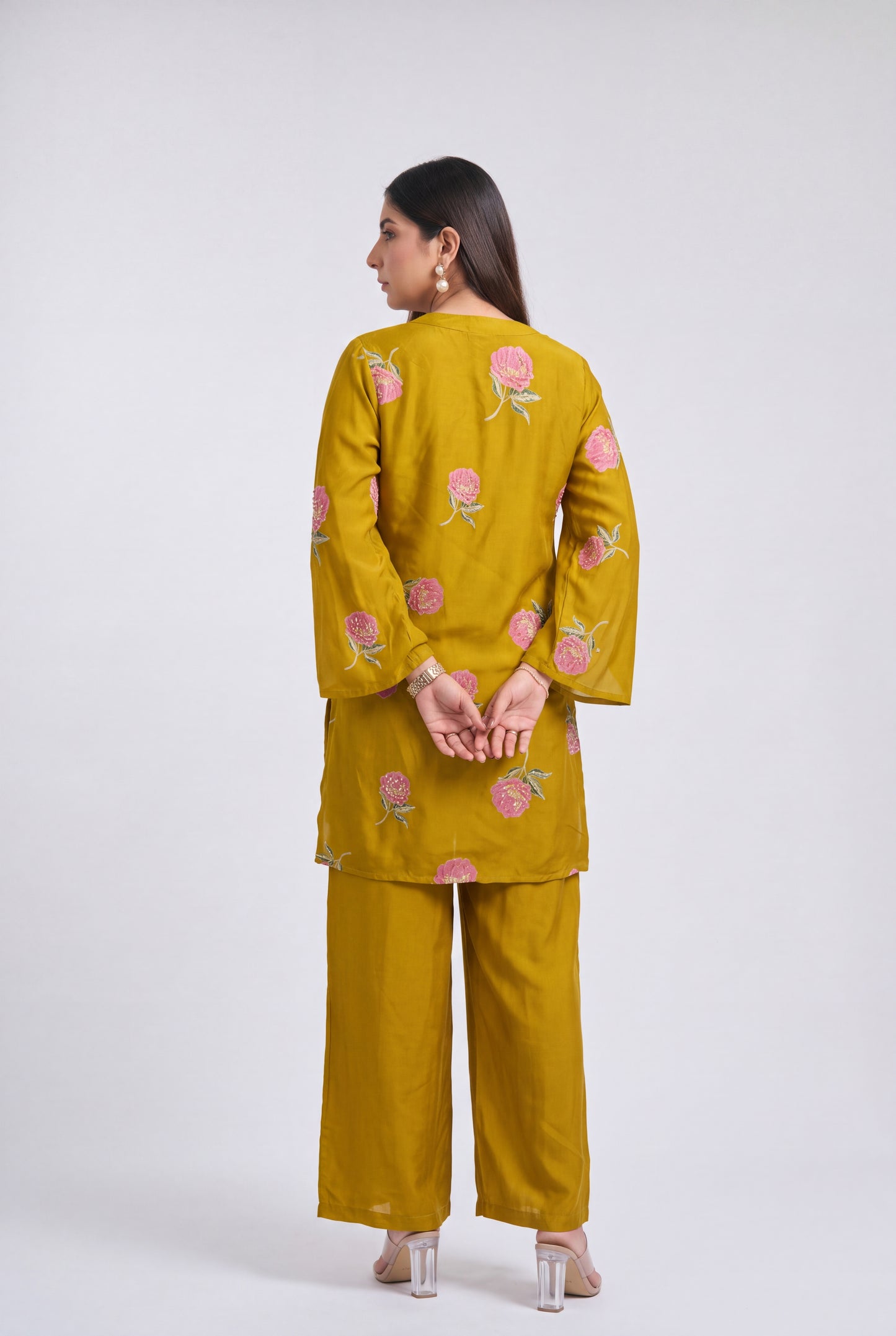 A woman is shown from the back wearing a mustard yellow kurta and matching wide-leg trousers, both decorated with pink floral embroidery, against a light gray studio background.