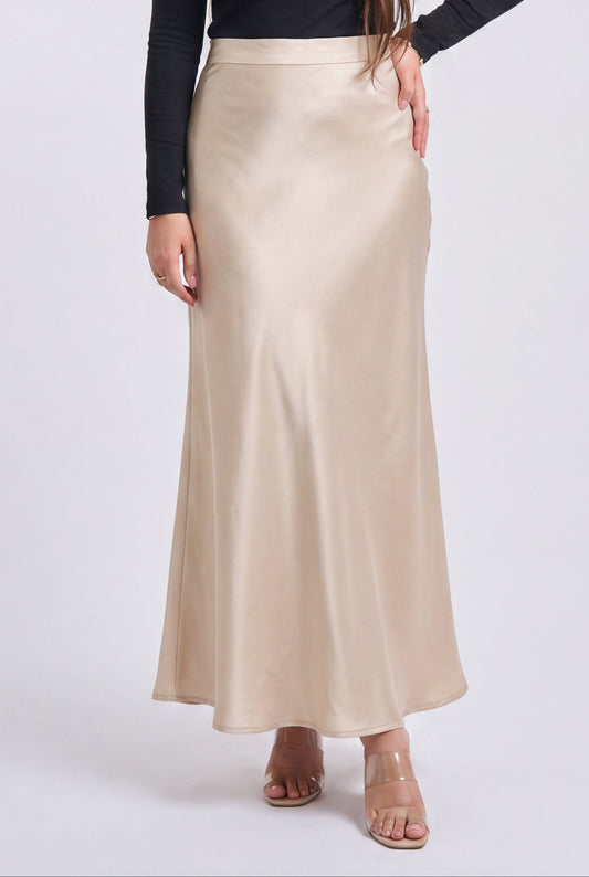A woman modeling a black long-sleeve top and a high-waisted, floor-length champagne beige satin skirt against a light gray studio background.