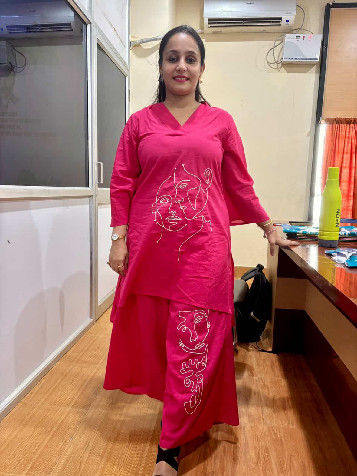 Woman in a pink dress standing in an indoor setting with a desk and window.