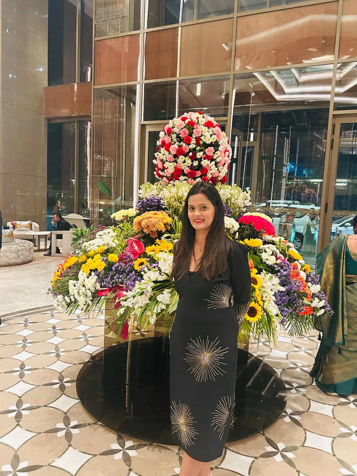 Woman standing next to a large floral arrangement in an indoor setting with reflective flooring.
