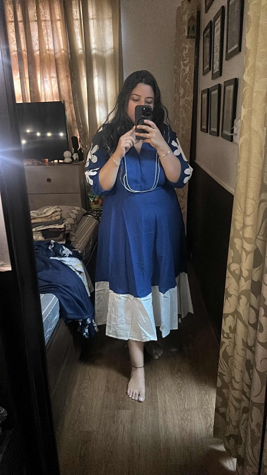 Woman taking a mirror selfie wearing a blue dress with white lace trim in a bedroom.
