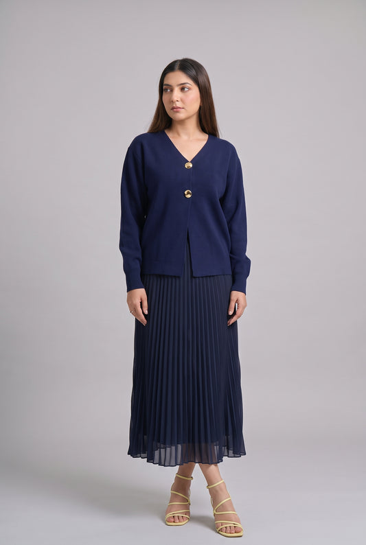 A woman modeling a navy blue cardigan with gold buttons and a matching pleated midi skirt, paired with beige strappy heels, against a solid gray studio background.