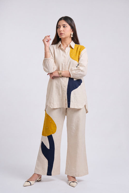 A woman modeling a matching two-piece outfit consisting of a cream-colored long-sleeve button-up shirt and wide-leg trousers, both decorated with abstract color-blocked patterns in mustard yellow, navy blue, and beige.