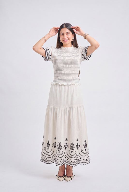 A woman modeling a cream-colored two-piece dress set featuring a smocked short-sleeve top and a tiered maxi skirt with black floral embroidery, set against a plain light gray studio background.