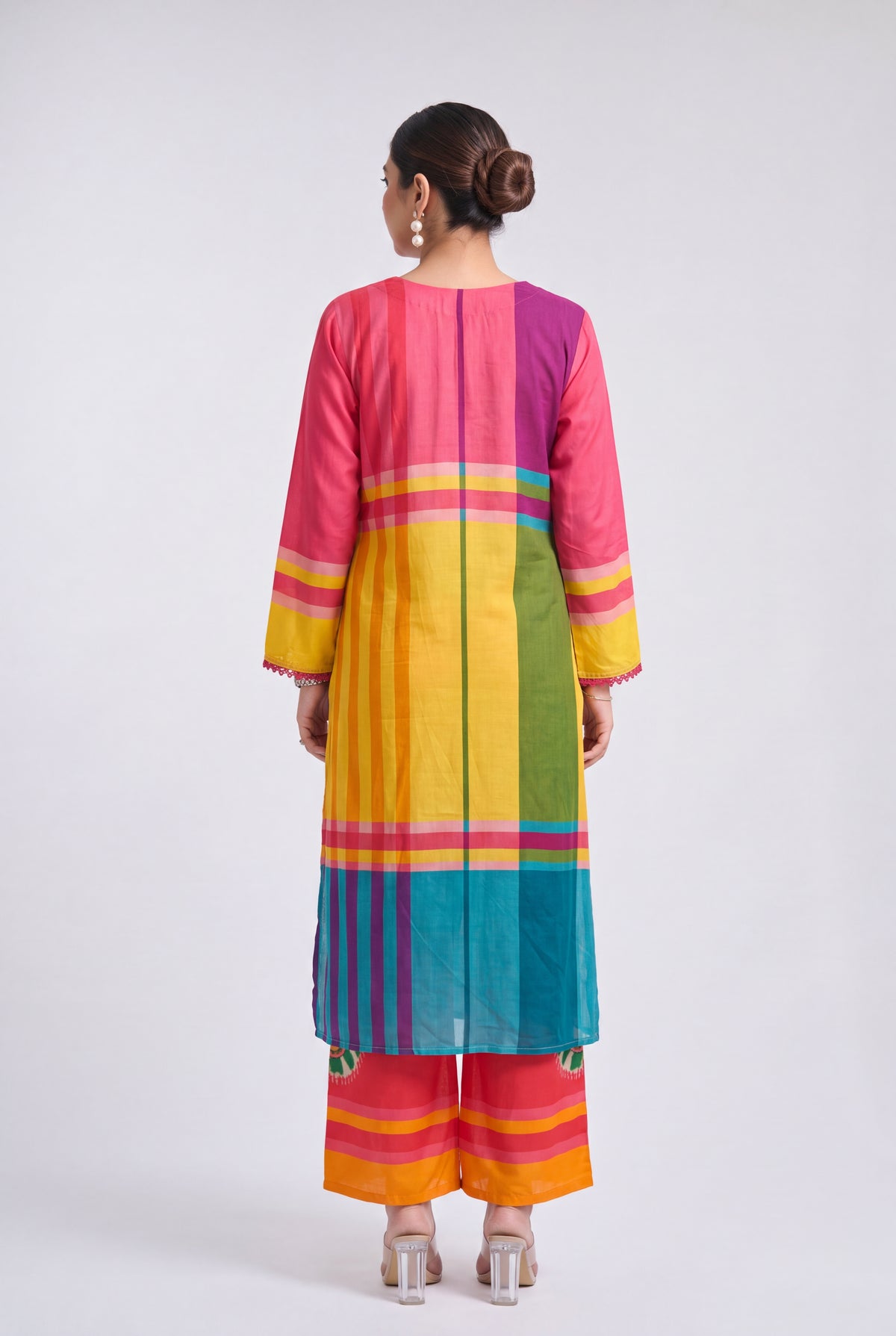 A woman viewed from the back wearing a brightly colored striped kurta and matching wide-leg pants. The outfit features vertical panels of pink, purple, yellow, green, blue, orange, and red with thin horizontal accent stripes, set against a plain light gray studio background.