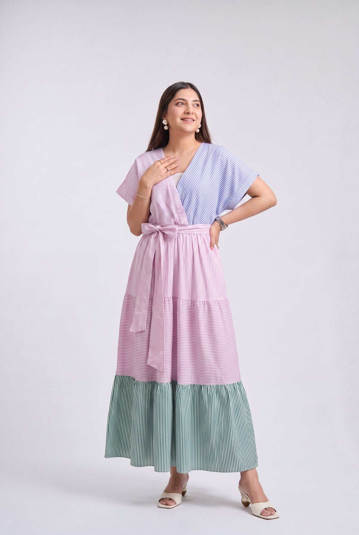 A woman modeling a three-tiered wrap maxi dress with a light purple upper section, a pink tiered skirt, and a sage green striped bottom, set against a plain light gray studio background.