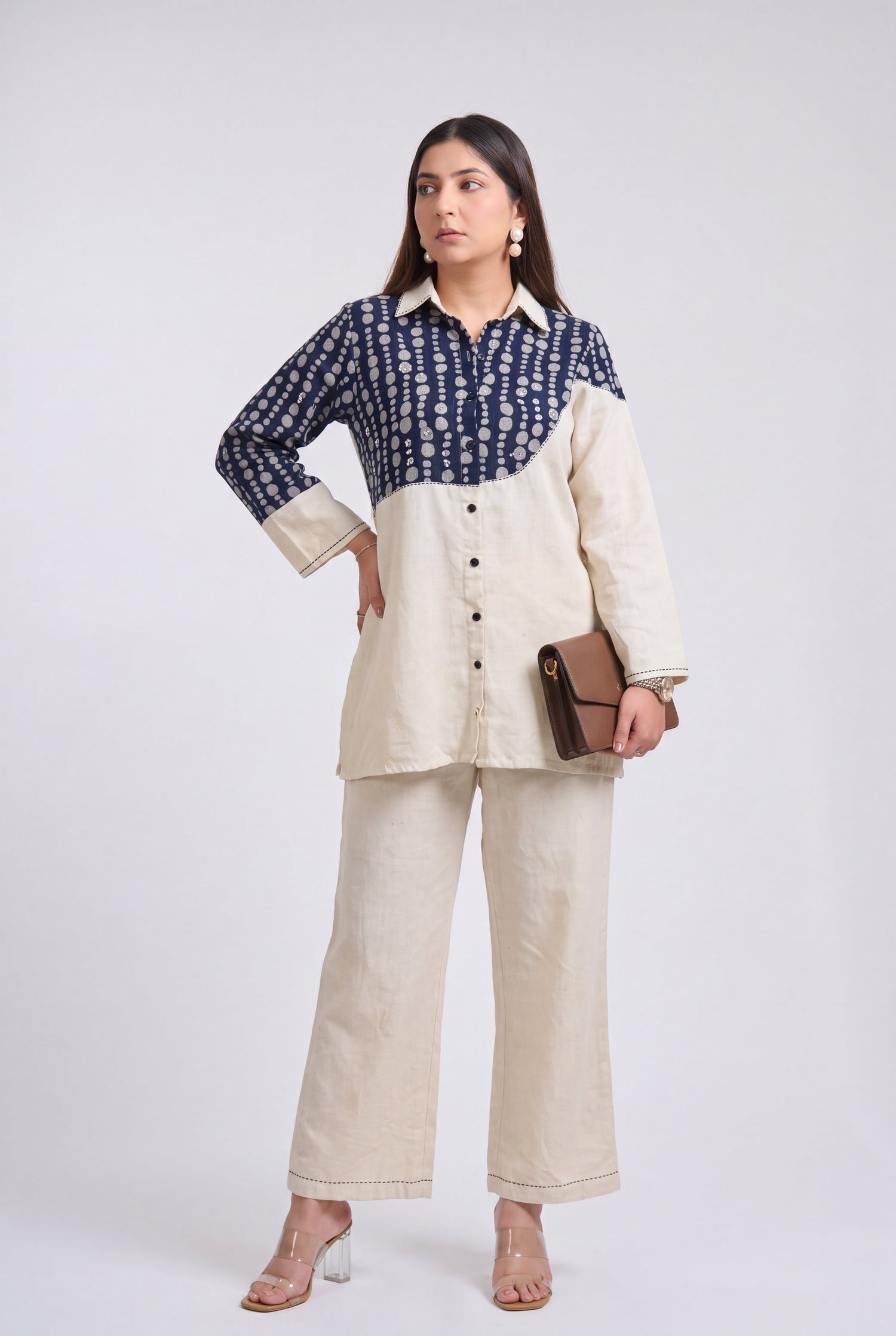 A woman models a two-tone shirt and trouser set against a plain light gray studio background. The shirt has a navy blue and cream patterned upper section and solid cream lower section, paired with matching cream wide-leg trousers.