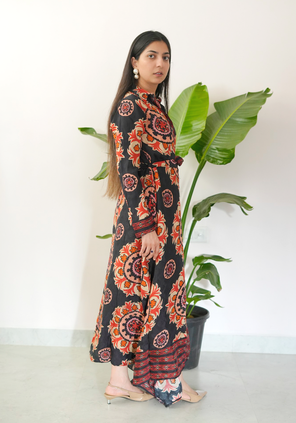 Woman in a patterned dress standing next to a plant against a white wall.