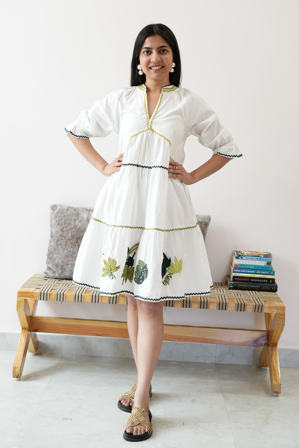 A woman standing on a bench, wearing a white knee-length dress with green and black embroidery, a V-neck, and quarter-length puffed sleeves.