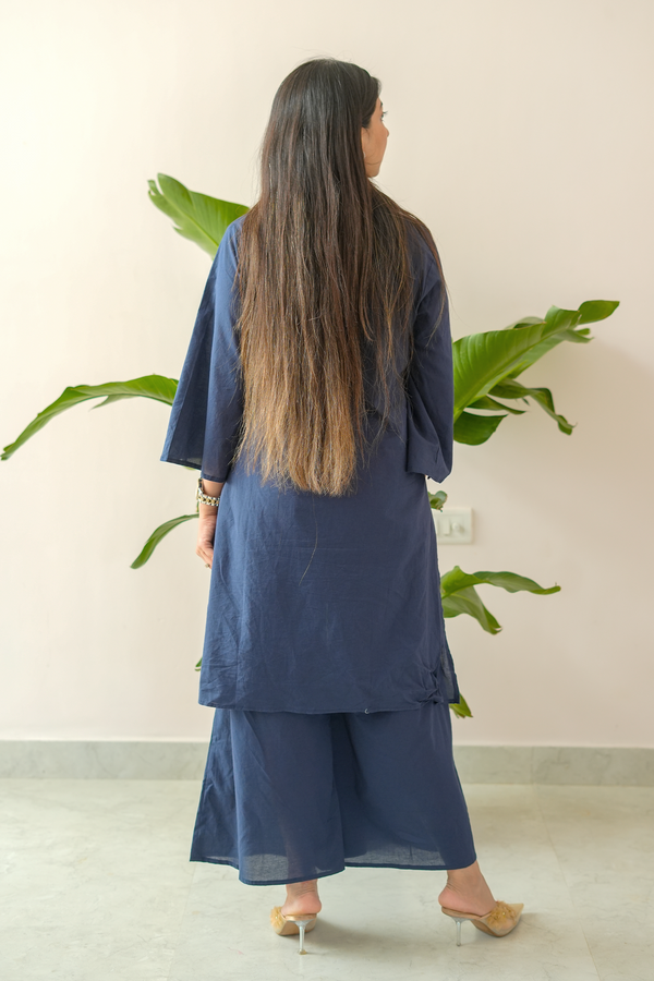 Woman wearing a blue outfit standing in front of a plant with a light pink wall.