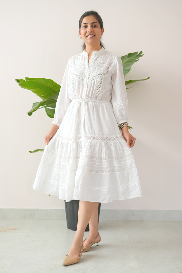 Woman wearing a white dress standing against a plain background with a plant.