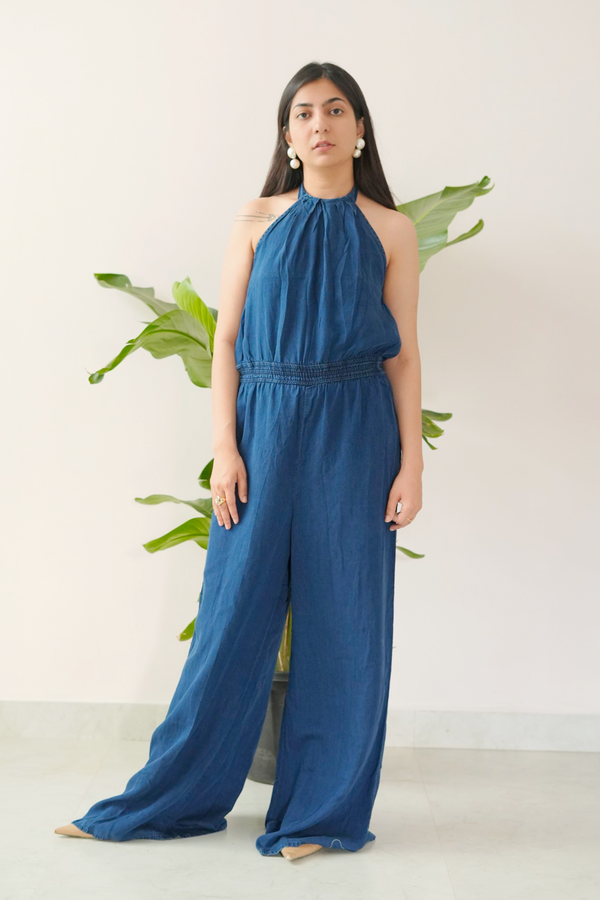 Woman wearing a blue jumpsuit standing against a white background with green plants.