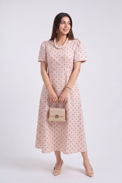 A woman modeling a light pink midi dress with small black polka dots, holding a beige handbag against a plain light gray studio background.