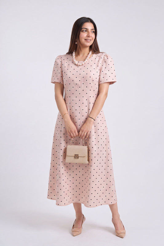 A woman modeling a light pink midi dress with small black polka dots, holding a beige handbag against a plain light gray studio background.