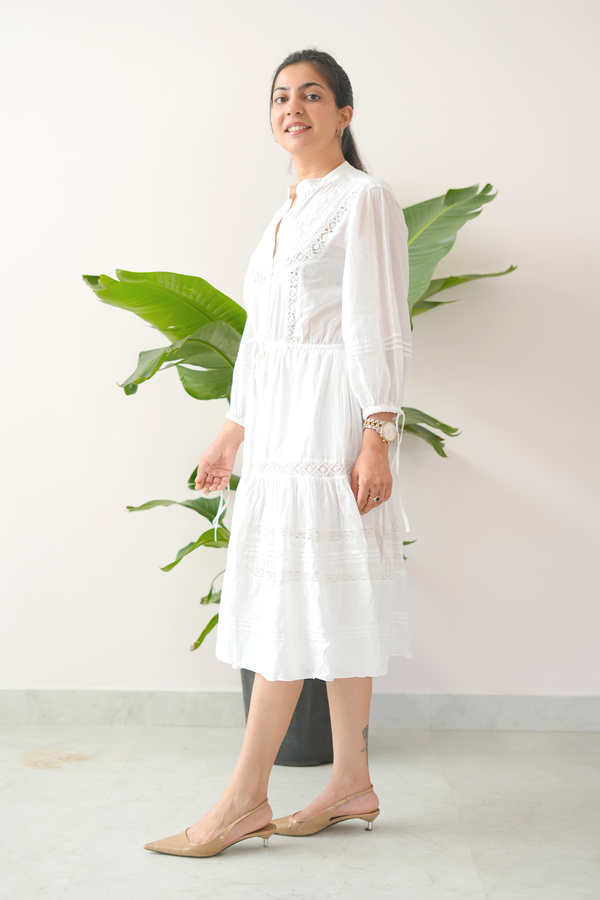 Woman wearing a white dress standing in front of a plant against a white wall.