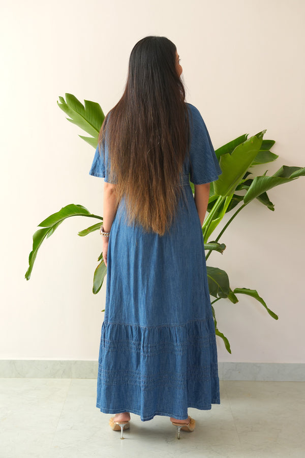 Woman wearing a blue dress standing in front of a large green plant against a plain wall.