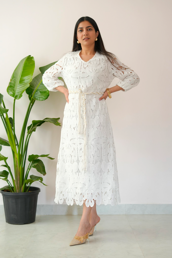 Woman wearing a white lace dress standing next to a potted plant against a plain wall.
