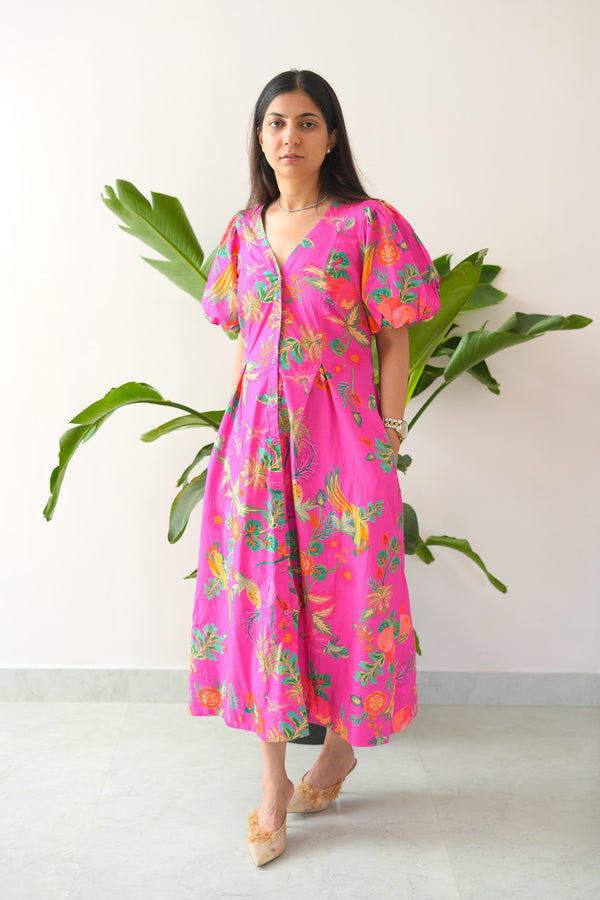 Woman wearing a bright pink floral dress standing next to a plant against a white wall.