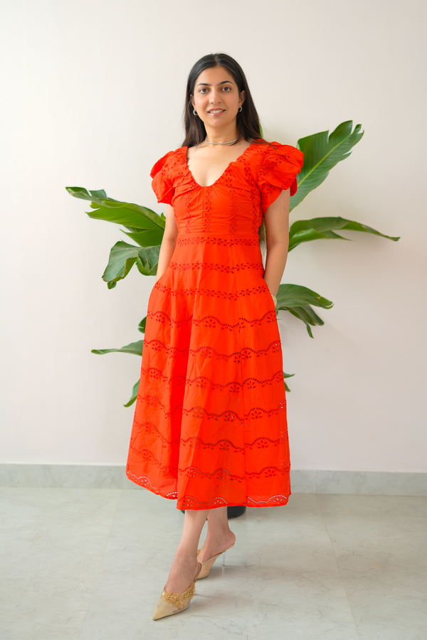 Woman wearing a bright orange dress with ruffled sleeves in a minimal indoor setting.