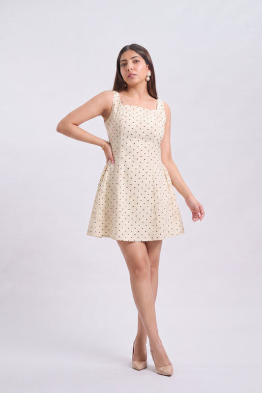 A woman modeling a sleeveless cream-colored mini dress with a small black polka dot pattern, standing against a light gray studio background.
