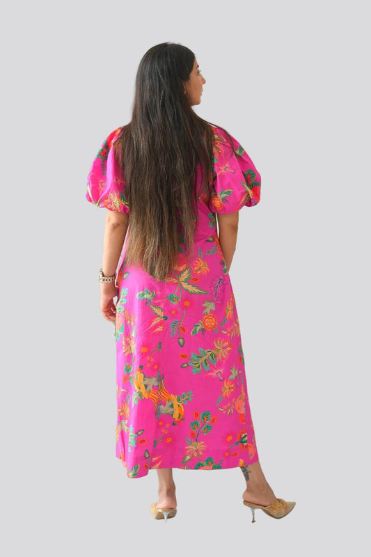 Woman wearing a bright pink floral dress standing next to a plant against a white wall.