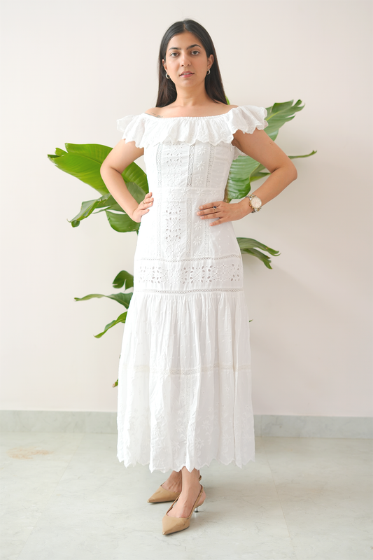 Woman wearing a white dress standing against a white wall with green plants.