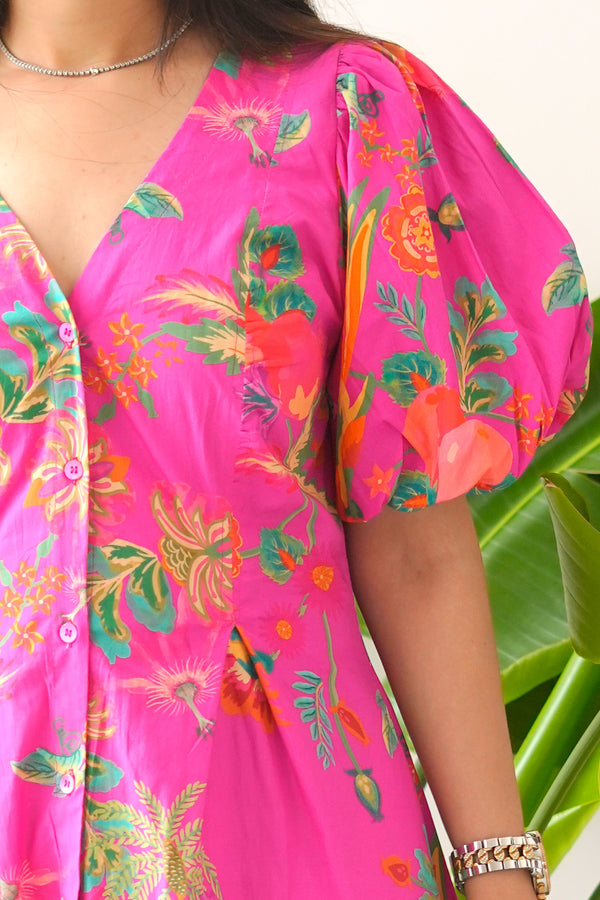 Bright pink dress with colorful floral pattern worn by a person, with a green plant in the background.
