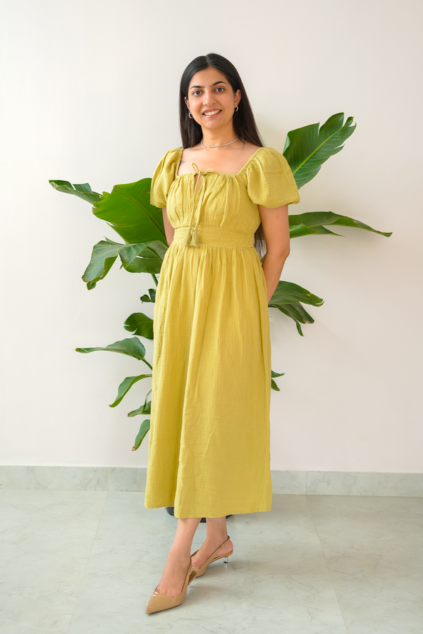 Woman in a yellow dress standing next to a plant against a white wall