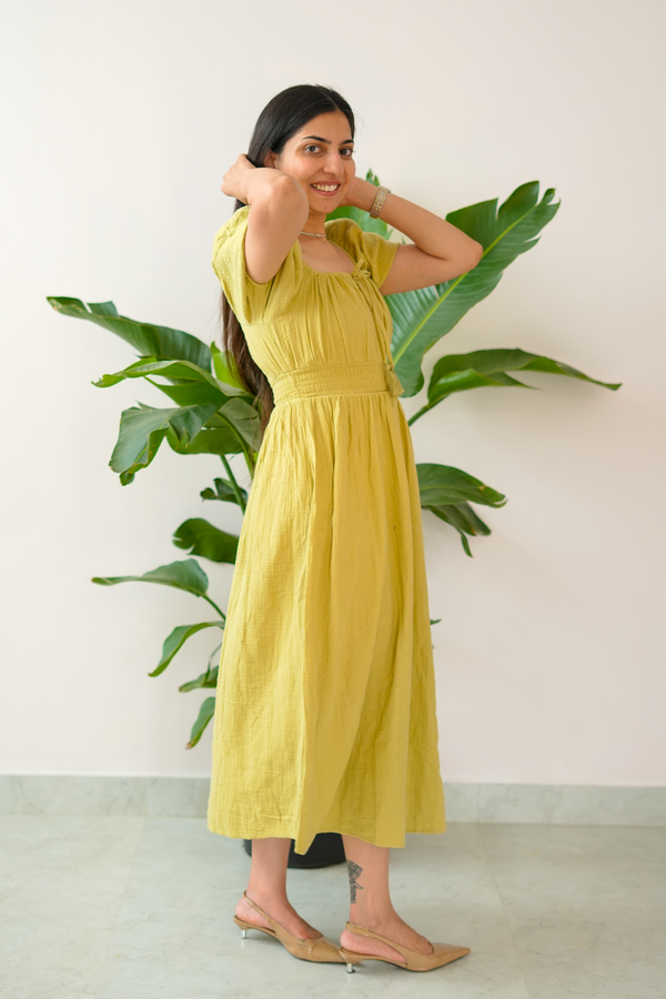 Woman in a yellow dress standing next to a plant against a white wall
