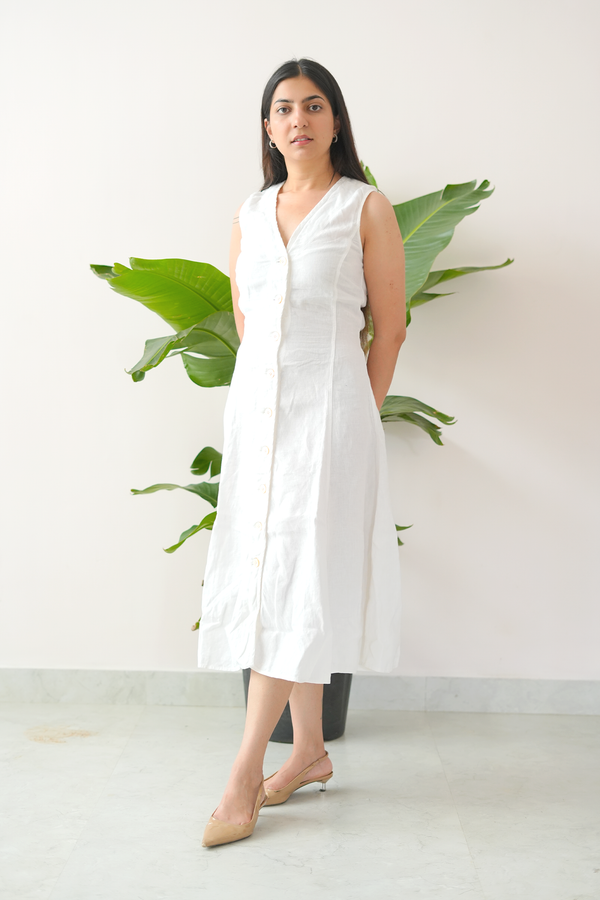 Woman wearing a white sleeveless dress standing in front of a plant against a white wall.