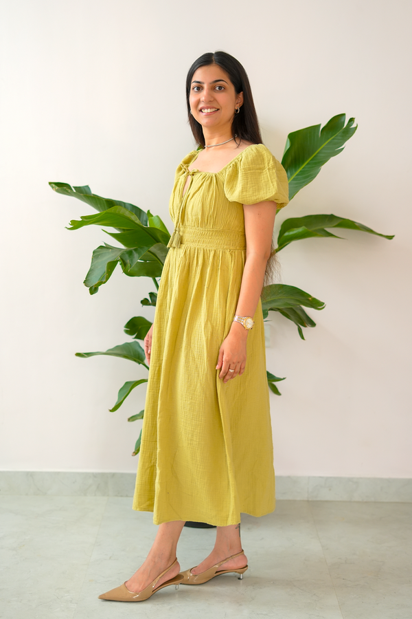 Woman wearing a yellow dress standing in front of a plant against a white wall.