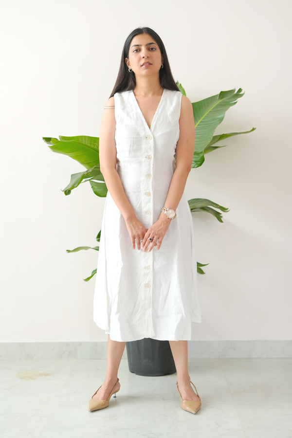 Woman wearing a white sleeveless dress standing against a white background with green plants.