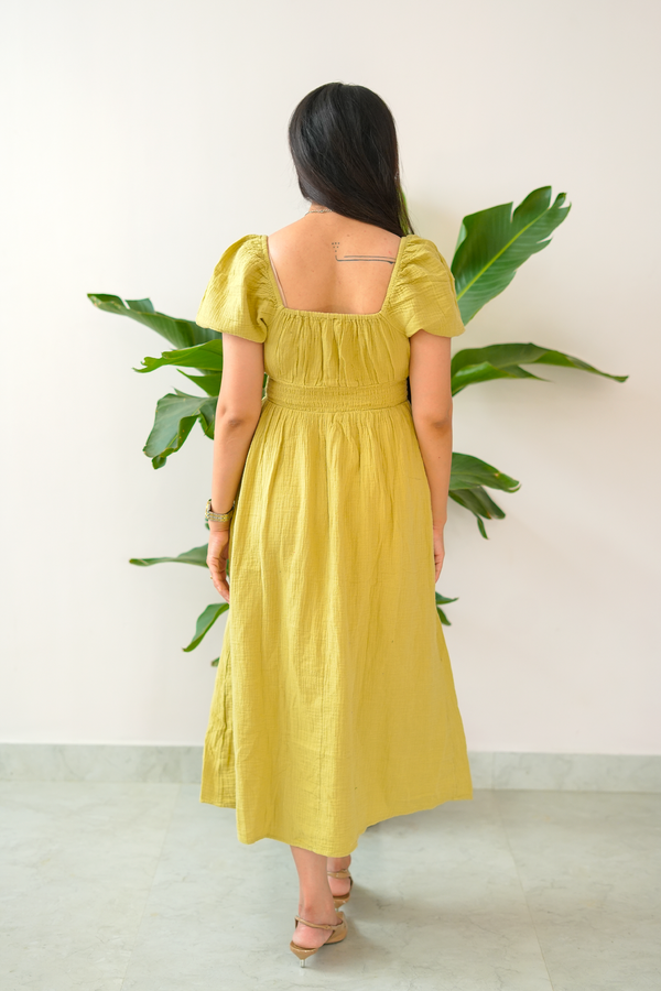 Woman wearing a yellow dress standing against a white wall with green plants.