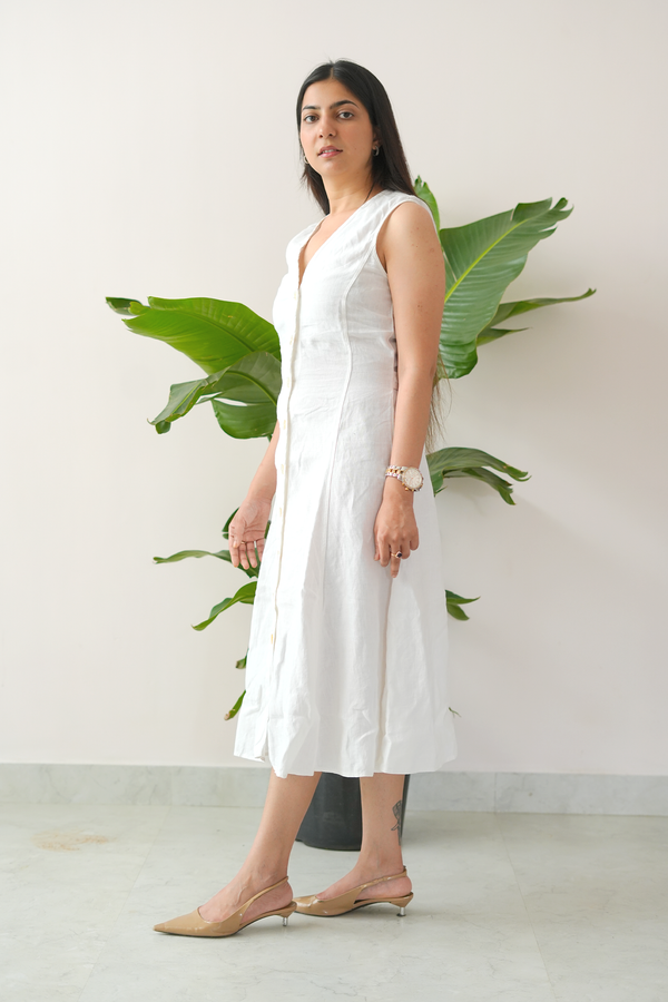 Woman in a white sleeveless dress standing against a white wall with green leaves.