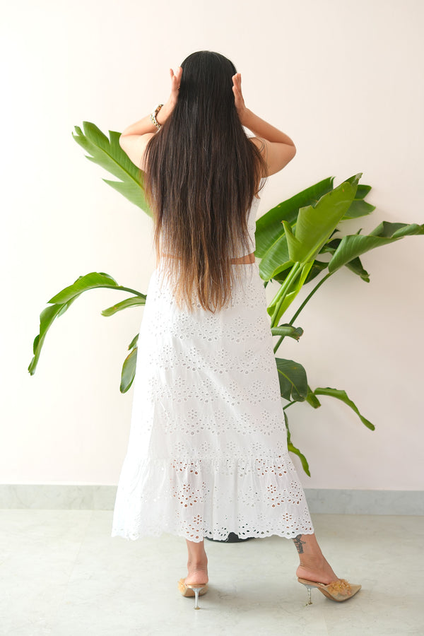 Woman in a white dress standing in front of a large green leafy plant against a white wall.