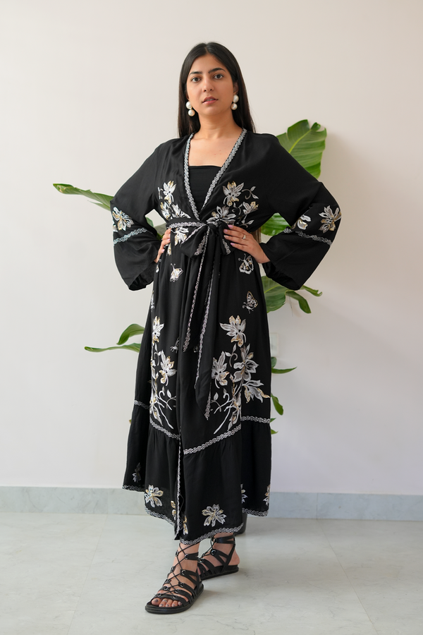 A woman standing in front of a plant backdrop wearing a long black dress with intricate floral embroidery, a belted waist, and wide sleeves.