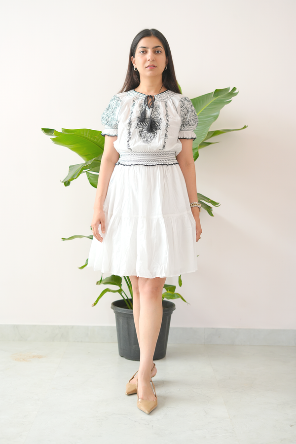 Woman wearing a white dress with black embroidery standing in front of a plant.