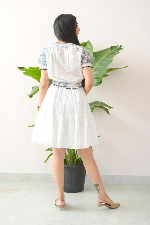 Woman wearing a white dress with embroidered details, standing in front of a white wall with green leaves.