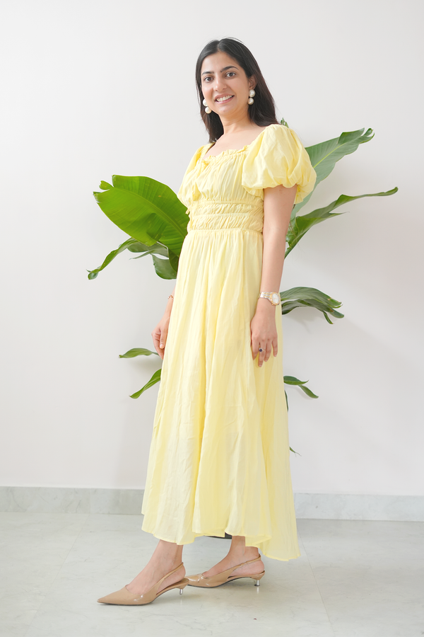 Woman wearing a yellow dress standing against a white wall with green leaves.
