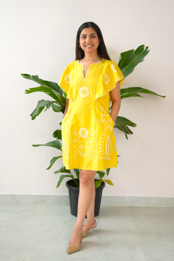 Woman wearing a yellow dress with white patterns standing next to a potted plant.