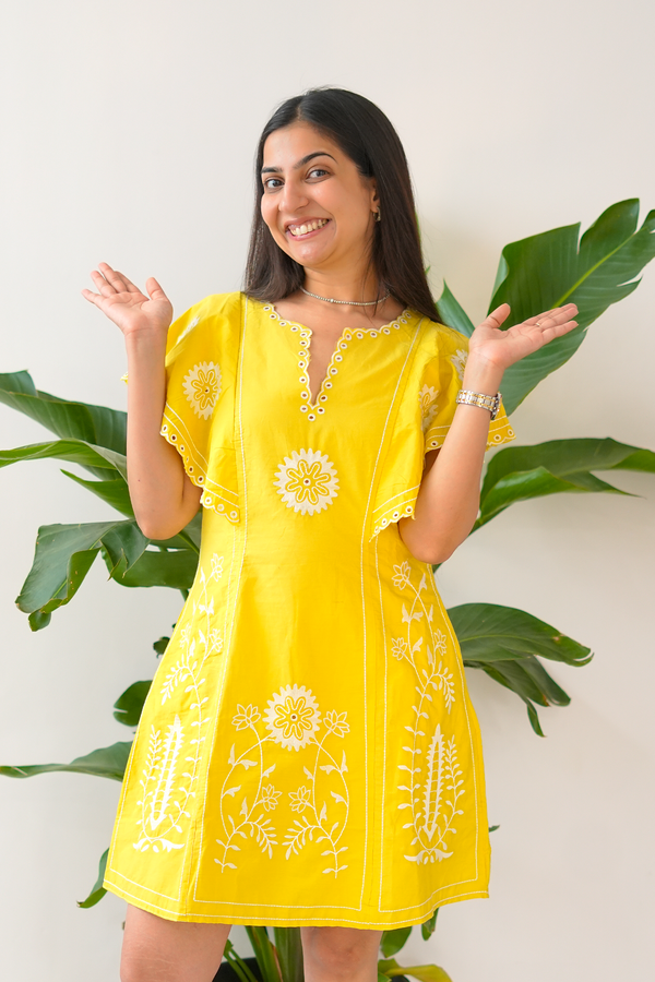 Woman wearing a yellow dress with floral patterns against a white wall with green leaves.