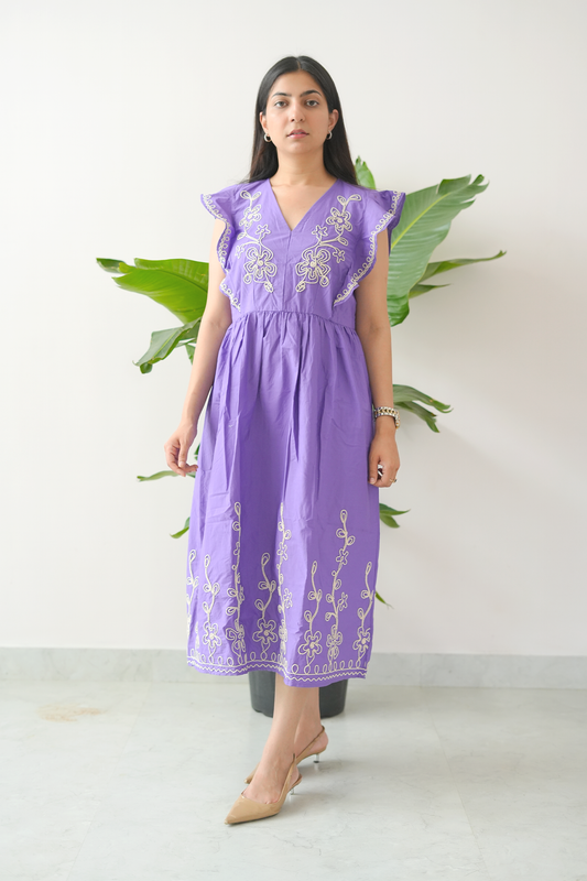 Woman wearing a purple embroidered dress standing against a white background with green plants.